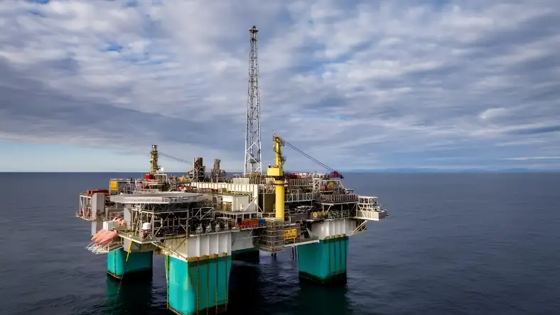 An offshore oil platform with yellow cranes and teal support legs standing in open sea under a cloudy sky