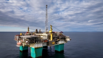 An offshore oil platform with yellow cranes and teal support legs standing in open sea under a cloudy sky